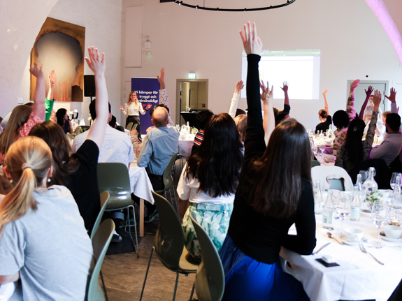 Bild från RKUF:s Riksårsmöte 2024. Unga personer är samlade vid bord och räcker upp sina händer för att visa på ungas organisering och engagemang.