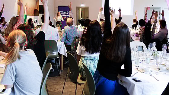 Bild från RKUF:s Riksårsmöte 2024. Unga personer sitter vid bord och räcker upp sina händer för att visa på ungas engagemang